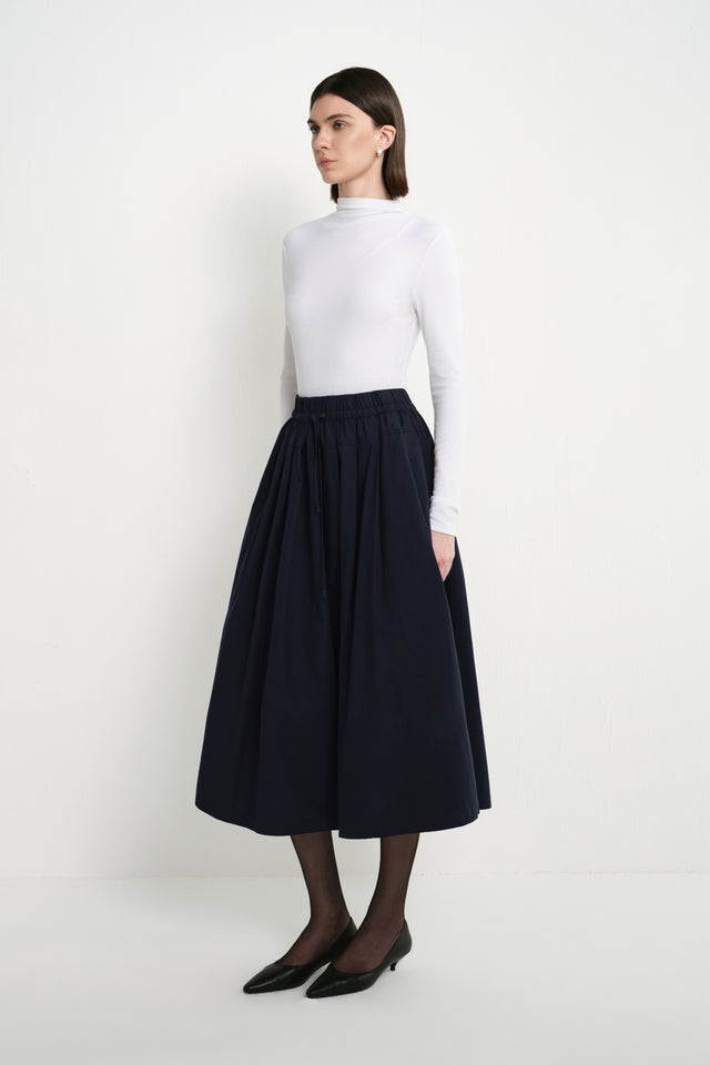 A model wearing the Rue Sophie Panier Drawstring Waist Pleated Skirt in Navy, showing its gathered volume and midi-length silhouette from a side angle.