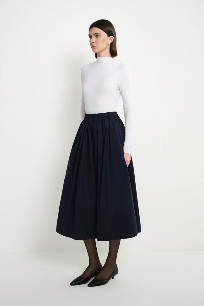 A model wearing the Rue Sophie Panier Drawstring Waist Pleated Skirt in Navy, showing its gathered volume and midi-length silhouette from a side angle.