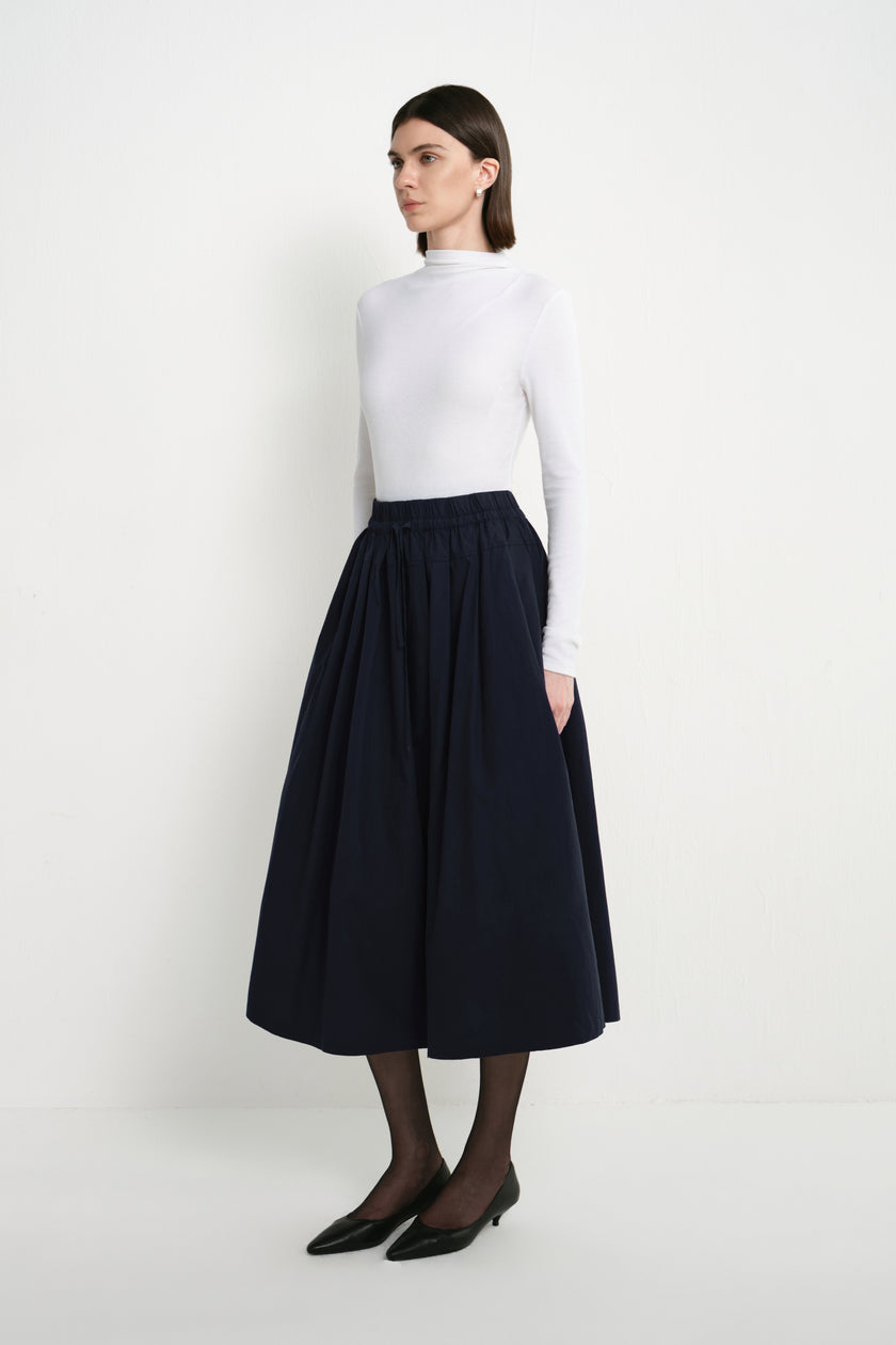 A model wearing the Rue Sophie Panier Drawstring Waist Pleated Skirt in Navy, showing its gathered volume and midi-length silhouette from a side angle.