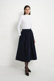 A model wearing the Rue Sophie Panier Drawstring Waist Pleated Skirt in Navy, styled with a white long-sleeve top and black heels, three-quarter front view.