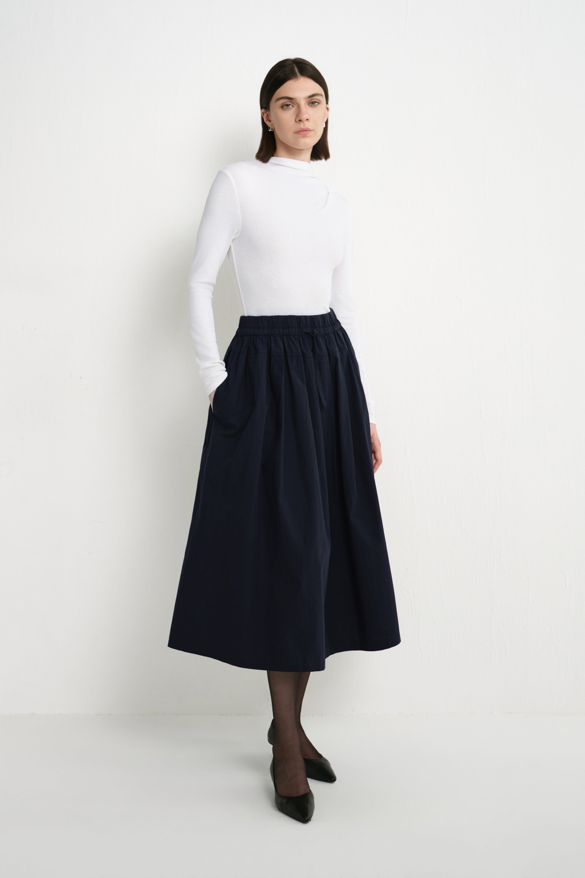 A model wearing the Rue Sophie Panier Drawstring Waist Pleated Skirt in Navy, styled with a white long-sleeve top and black heels, three-quarter front view.
