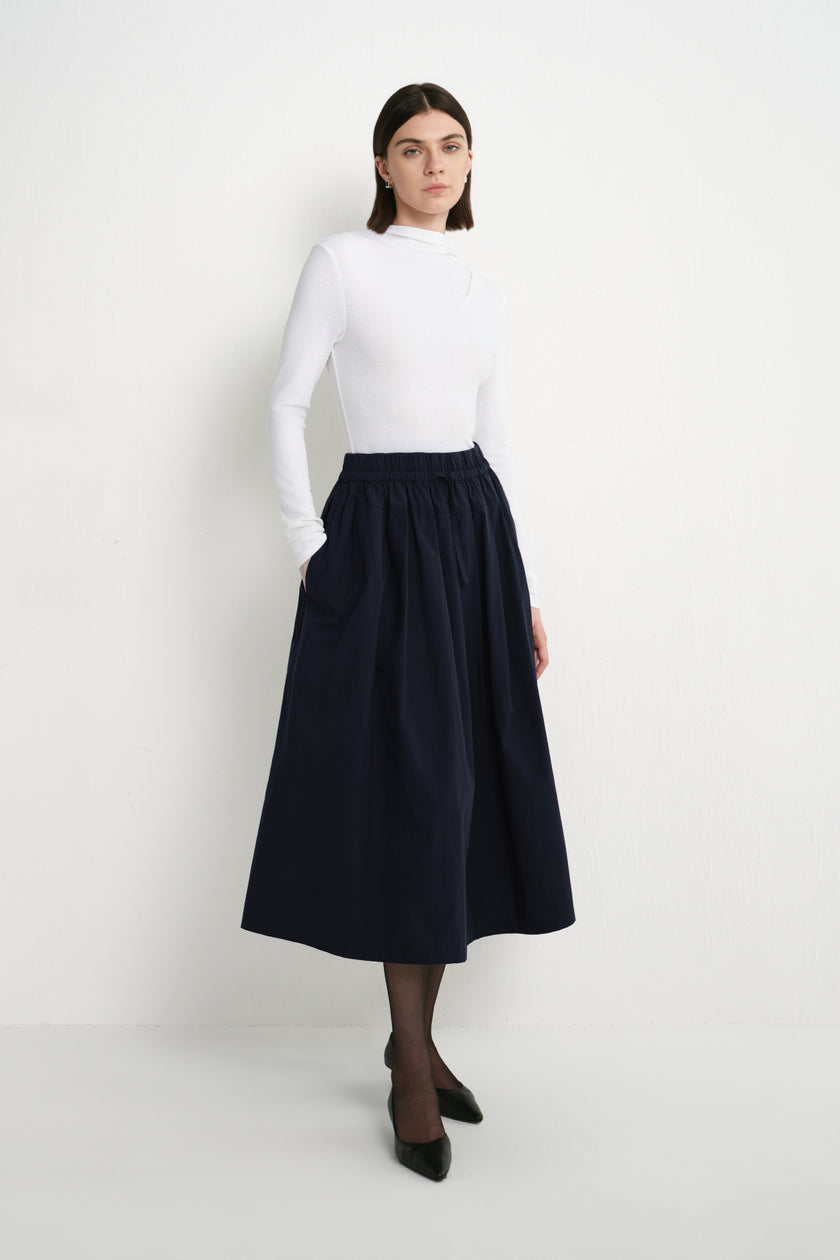 A model wearing the Rue Sophie Panier Drawstring Waist Pleated Skirt in Navy, styled with a white long-sleeve top and black heels, three-quarter front view.
