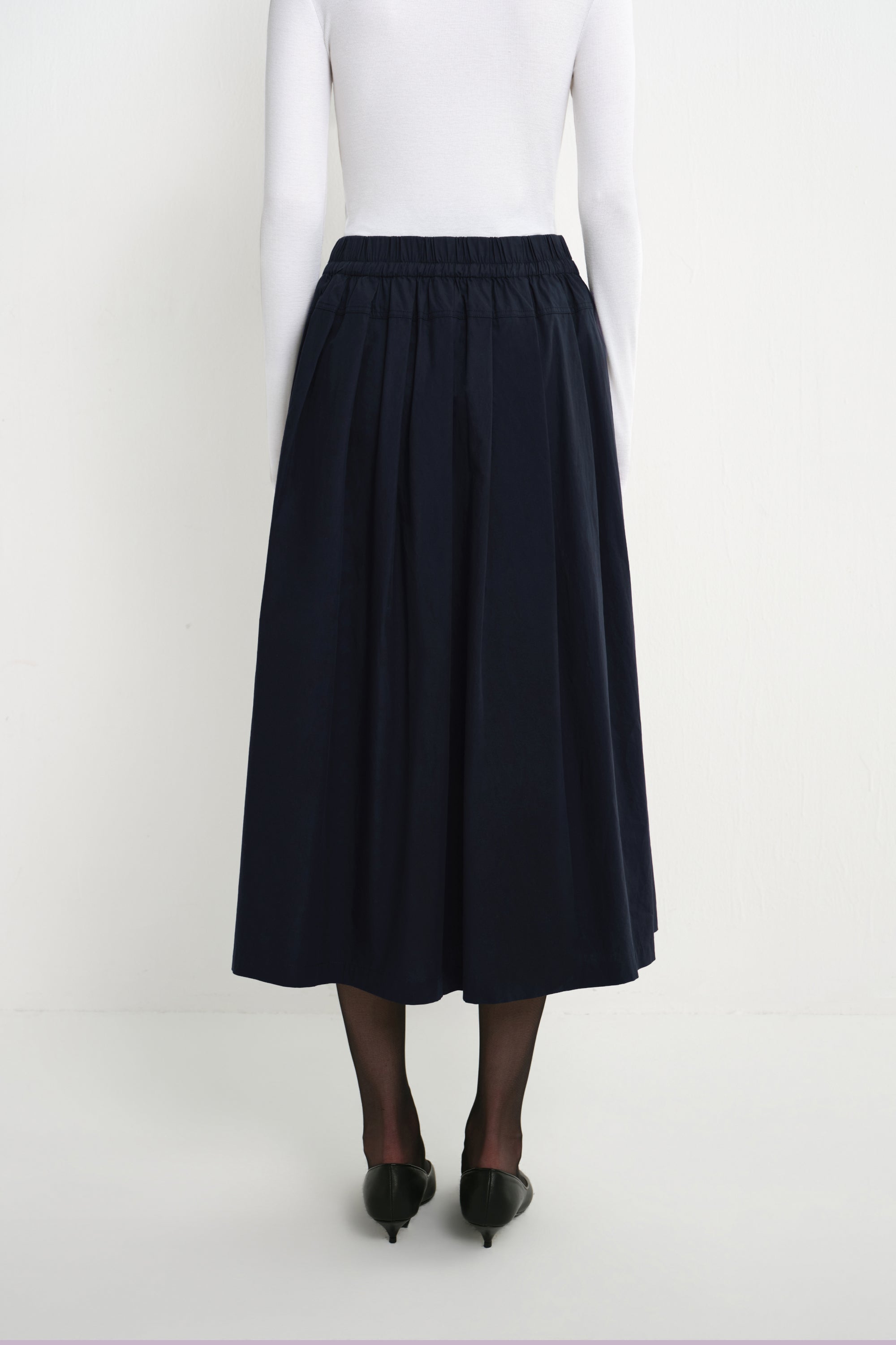 Back view of a model wearing the Rue Sophie Panier Drawstring Waist Pleated Skirt in Navy, showing the elasticated waist and full gathered silhouette.