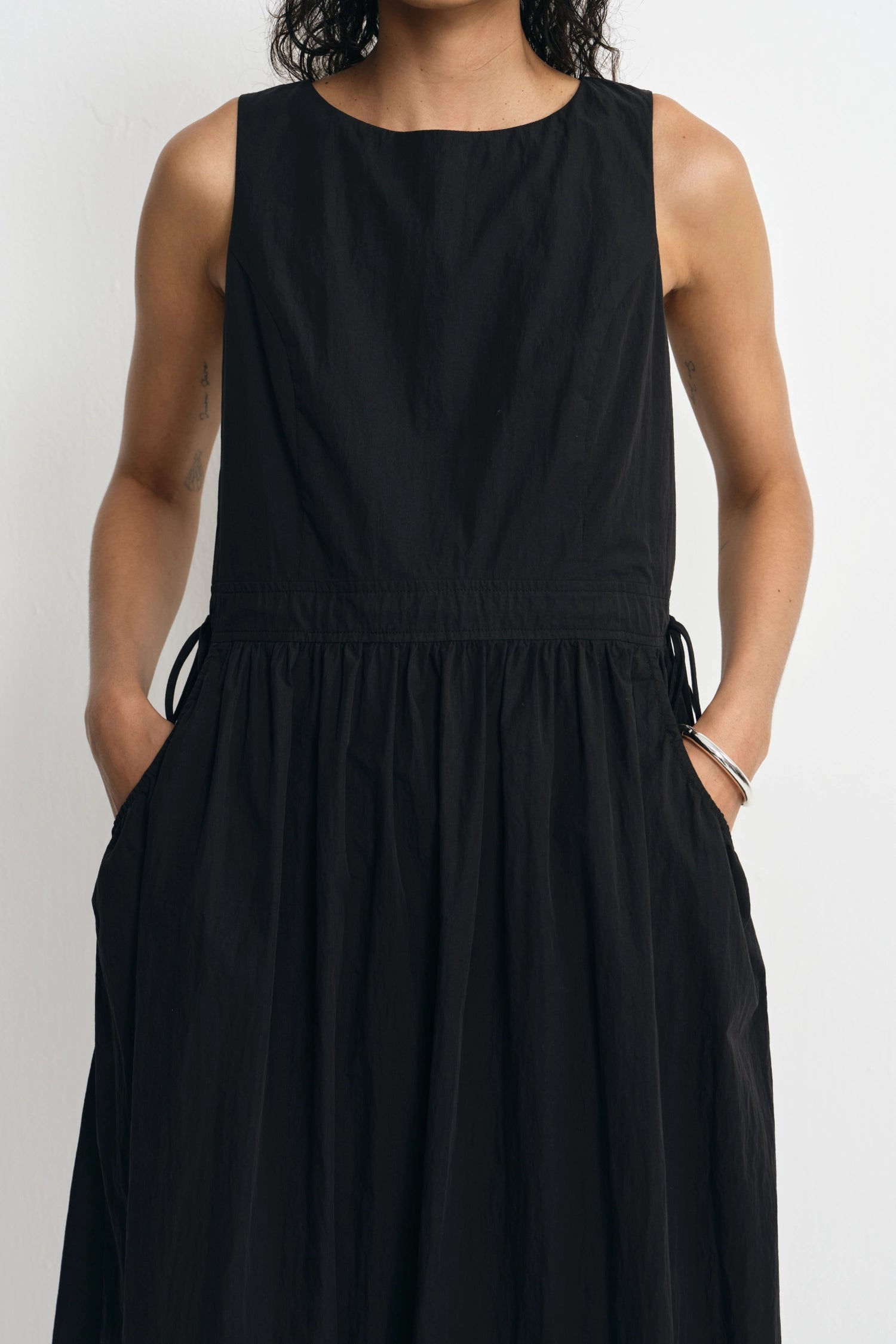 A sleeveless black dress with a round neckline, gathered waist, side drawstrings, and side pockets, shown on a model against a light gray background.