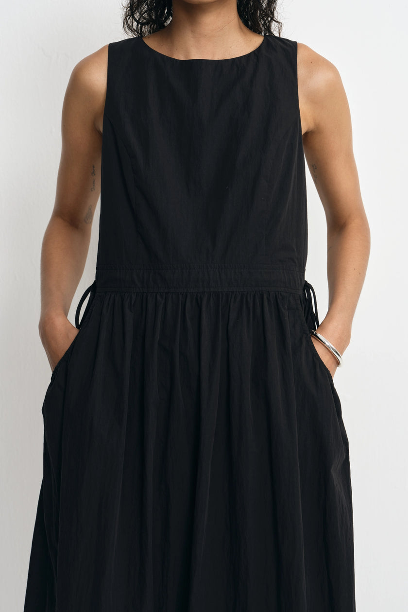 A sleeveless black dress with a round neckline, gathered waist, side drawstrings, and side pockets, shown on a model against a light gray background.