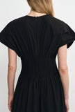 Back view of a woman wearing a black pleated dress with short wide sleeves and a gathered waist, shown against a plain light background.