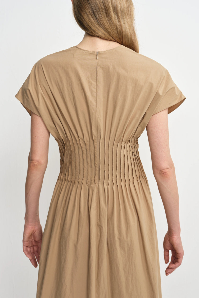 A woman viewed from the back wearing a short-sleeve beige dress with a gathered smocked waist and a small back zipper, photographed against a plain light gray studio background.