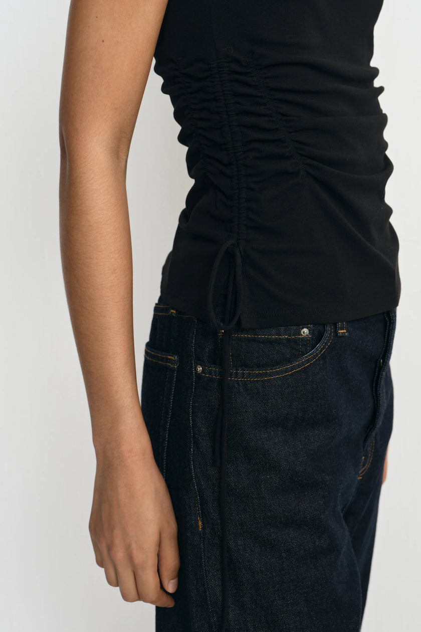 Close-up of a person wearing a black ruched sleeveless top and dark blue jeans against a light gray background.