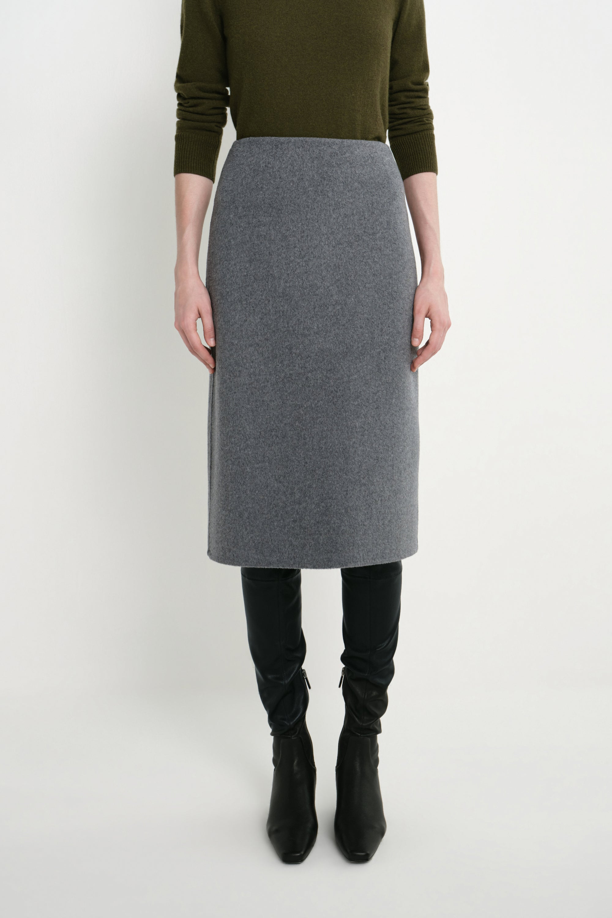 A model wearing a gray wool-blend pencil skirt, styled with an olive green long-sleeve top, black leggings, and black ankle boots, photographed against a light gray studio background.