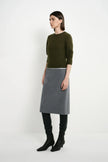 A model stands in profile wearing a dark olive green long-sleeve knit top, a grey knee-length skirt, black leggings, and black heeled boots against a plain white studio background.