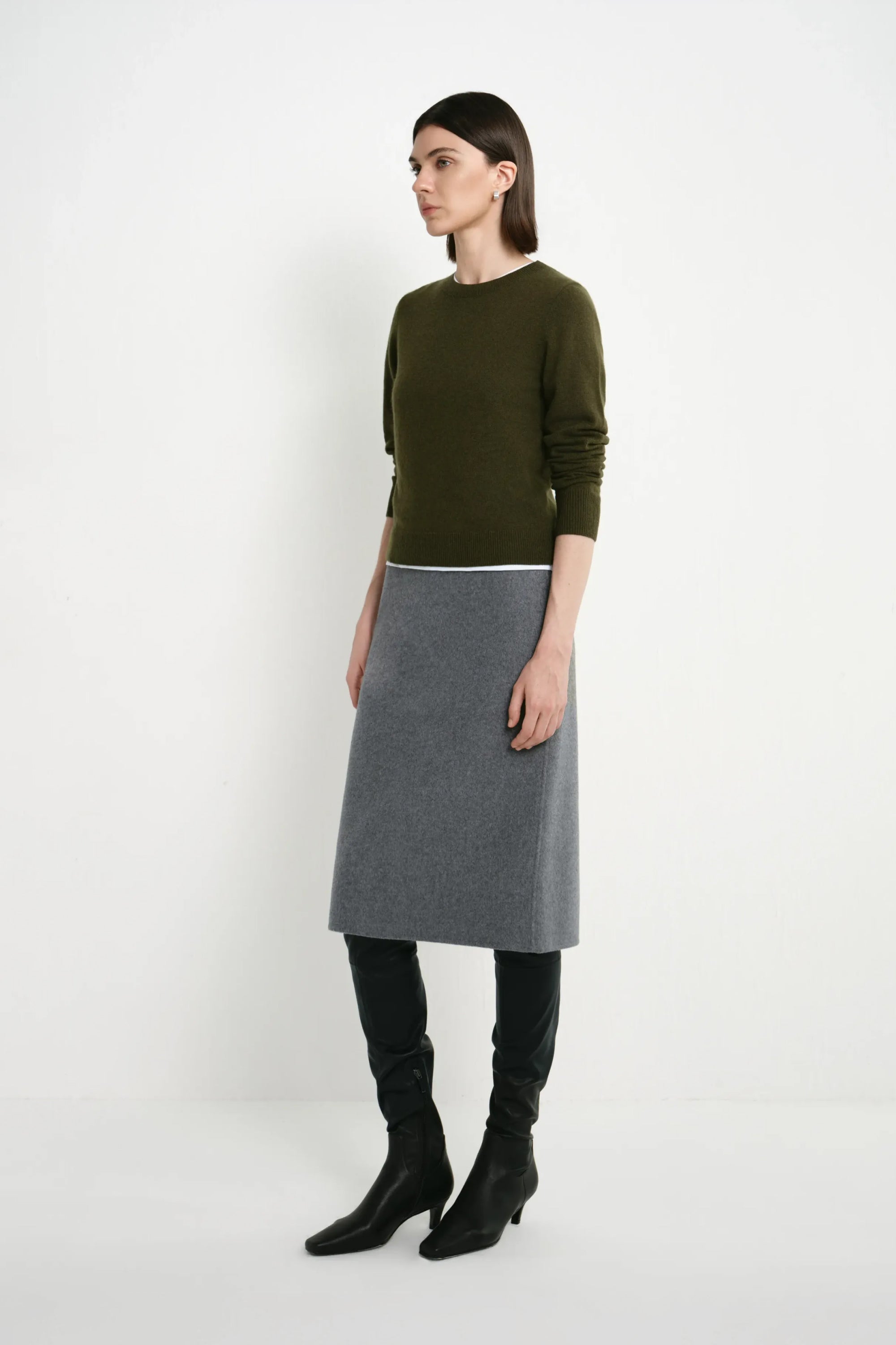 A model stands in profile wearing a dark olive green long-sleeve knit top, a grey knee-length skirt, black leggings, and black heeled boots against a plain white studio background.