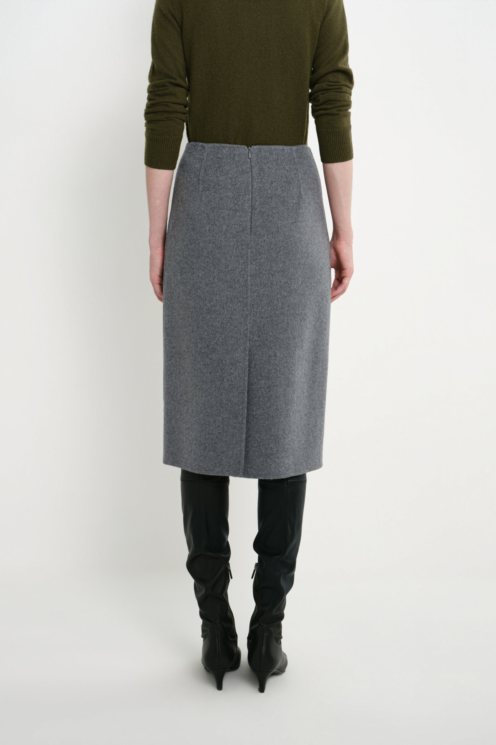 A model shown from the back wearing a gray wool-blend midi skirt, an olive green sweater, and black knee-high boots against a plain white studio background.