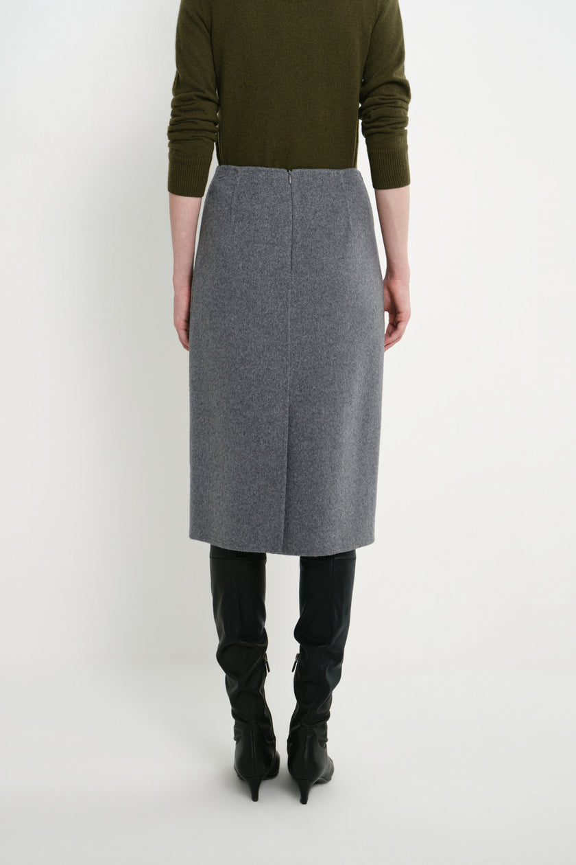 A model shown from the back wearing a gray wool-blend midi skirt, an olive green sweater, and black knee-high boots against a plain white studio background.