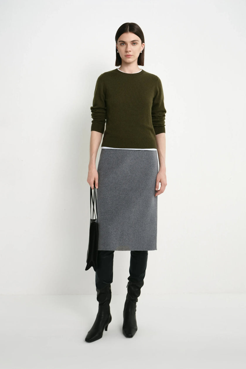 A model stands against a plain light gray studio background wearing a color-blocked sweater and skirt set. The top is olive green, the middle band is white, and the skirt is heather gray.