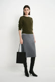 A model stands against a light gray studio background wearing an olive green long-sleeve sweater, a gray knee-length skirt, black over-the-knee boots, and holding a black tote bag.