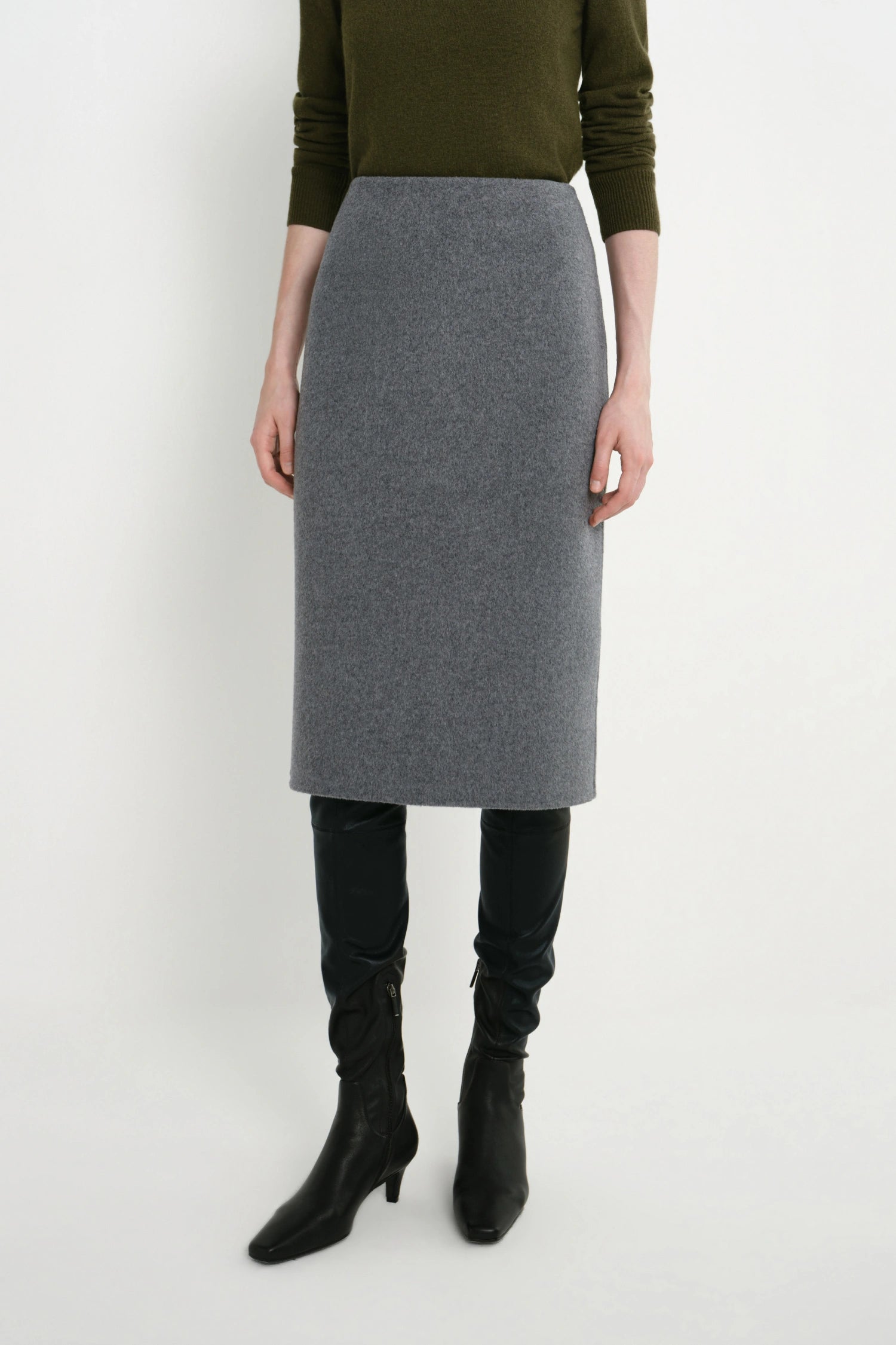 A model wearing a gray knit pencil skirt, an olive green long-sleeve top, black over-the-knee boots, and black pointed-toe heeled ankle boots, standing against a plain white studio background.