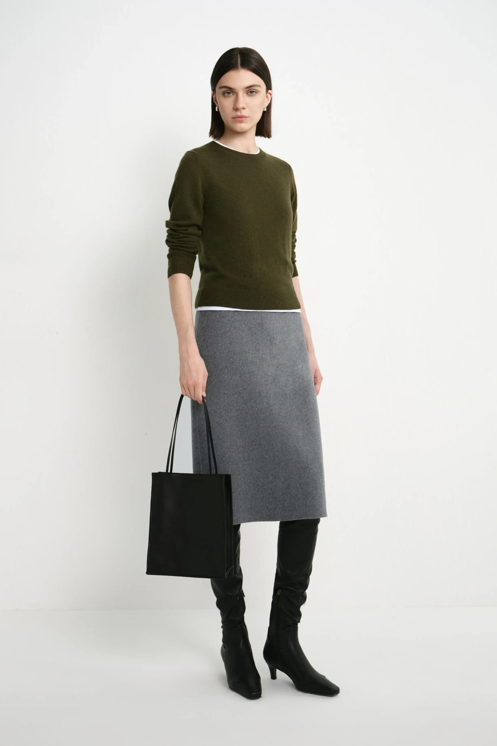 A model stands against a light gray studio background wearing an olive green long-sleeve sweater, a gray knee-length skirt, black over-the-knee boots, and holding a black tote bag.