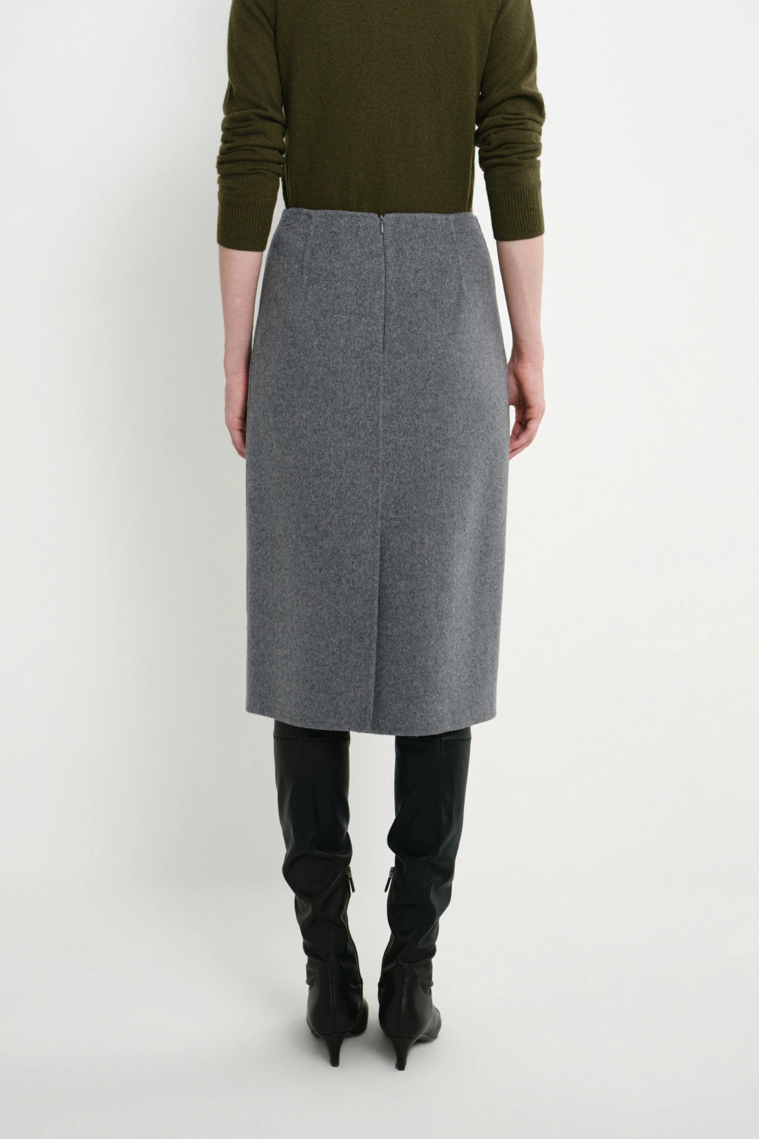 A model shown from the back wearing a gray wool-blend midi skirt, an olive green sweater, and black knee-high boots against a plain white studio background.
