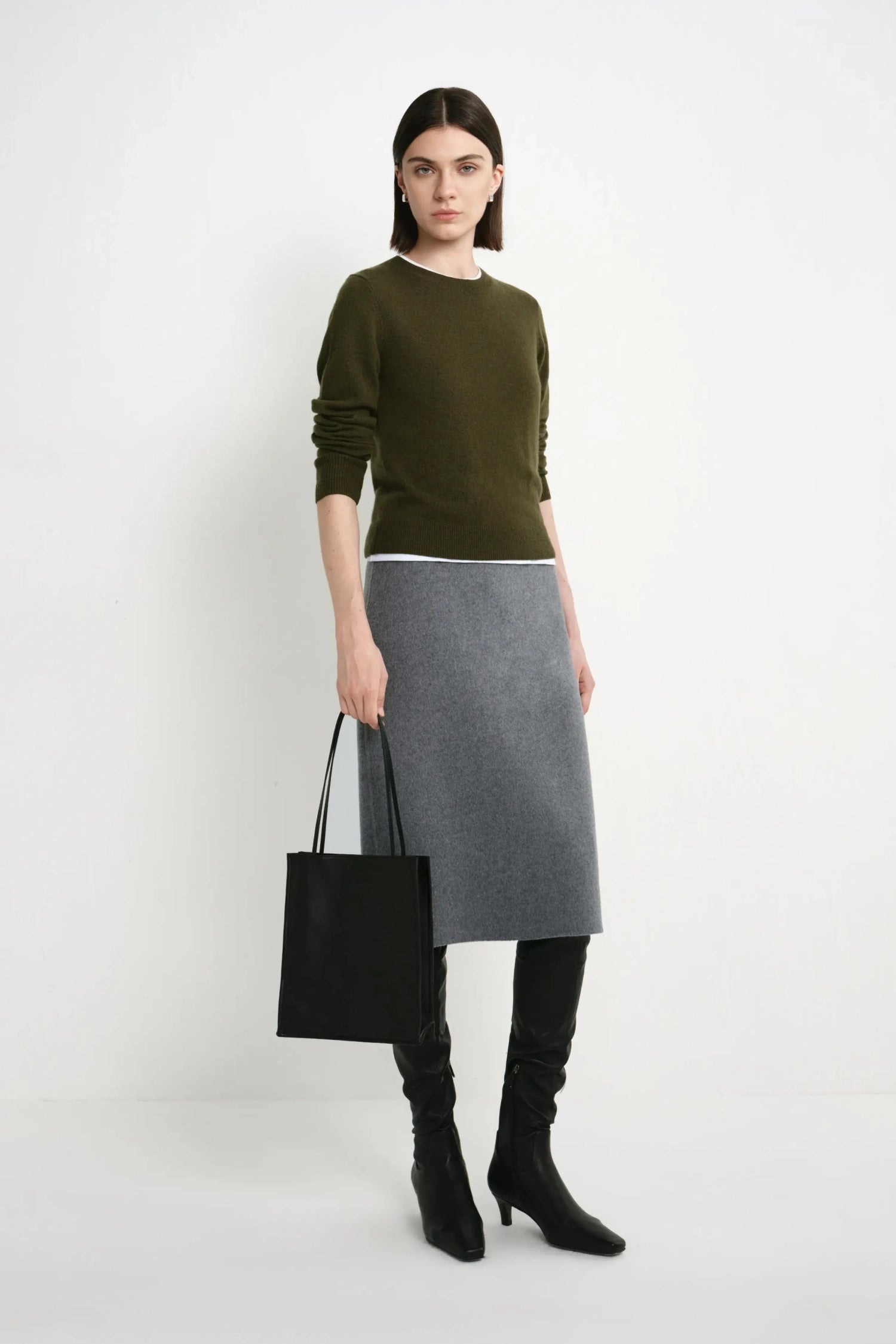 A model stands against a light gray studio background wearing an olive green long-sleeve sweater, a gray knee-length skirt, black over-the-knee boots, and holding a black tote bag.