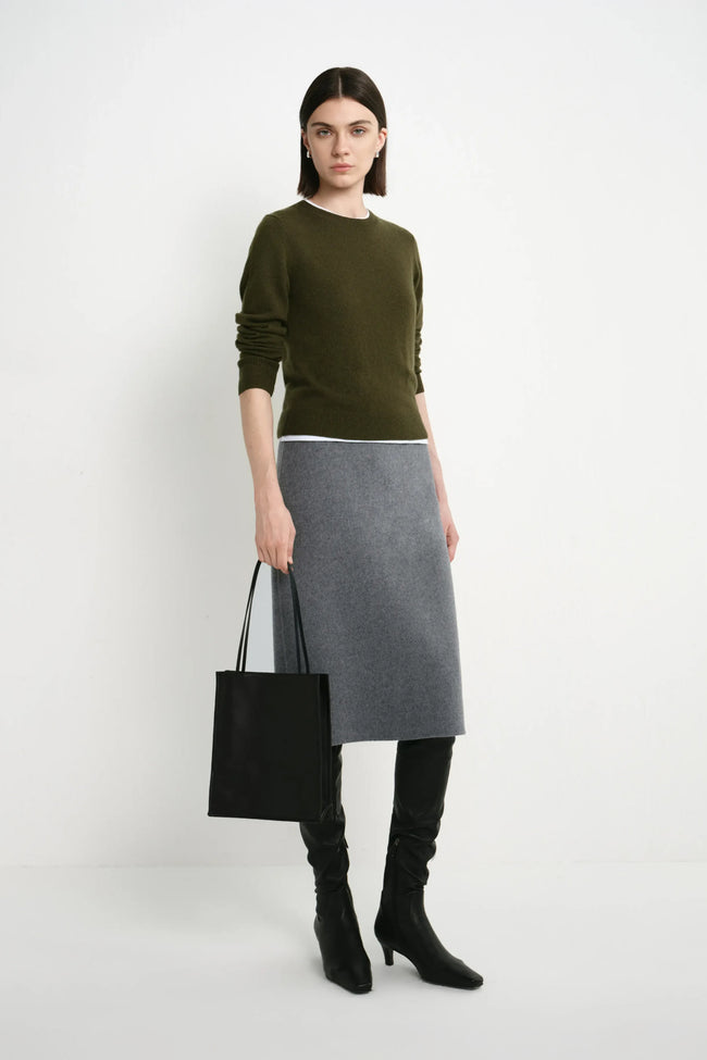 A model stands against a light gray studio background wearing an olive green long-sleeve sweater, a gray knee-length skirt, black over-the-knee boots, and holding a black tote bag.