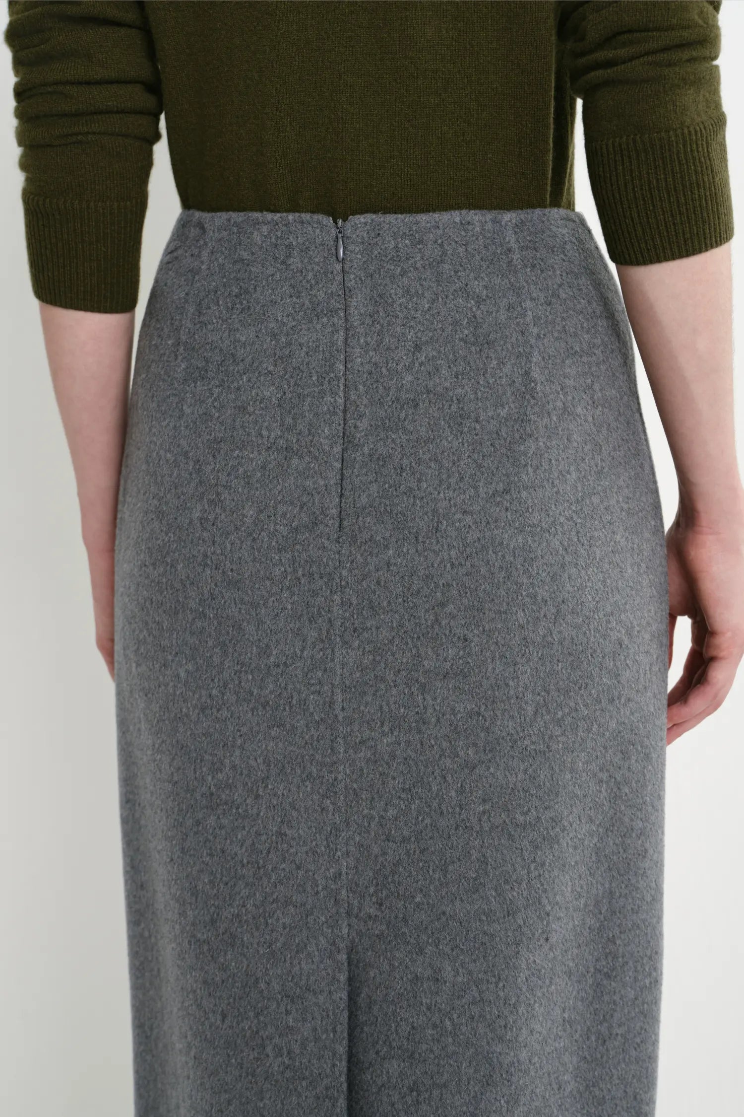A close-up of a person wearing a high-waisted, wide-leg gray wool skirt paired with an olive green ribbed knit top, shown from the back against a plain light background.