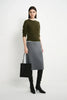 A model stands against a light gray studio background wearing an olive green long-sleeve sweater, a gray knee-length skirt, black over-the-knee boots, and holding a black tote bag.