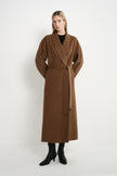 A woman modeling a long brown wrap coat with a tie waist, worn over a black top and black ankle boots, standing against a plain white studio background.