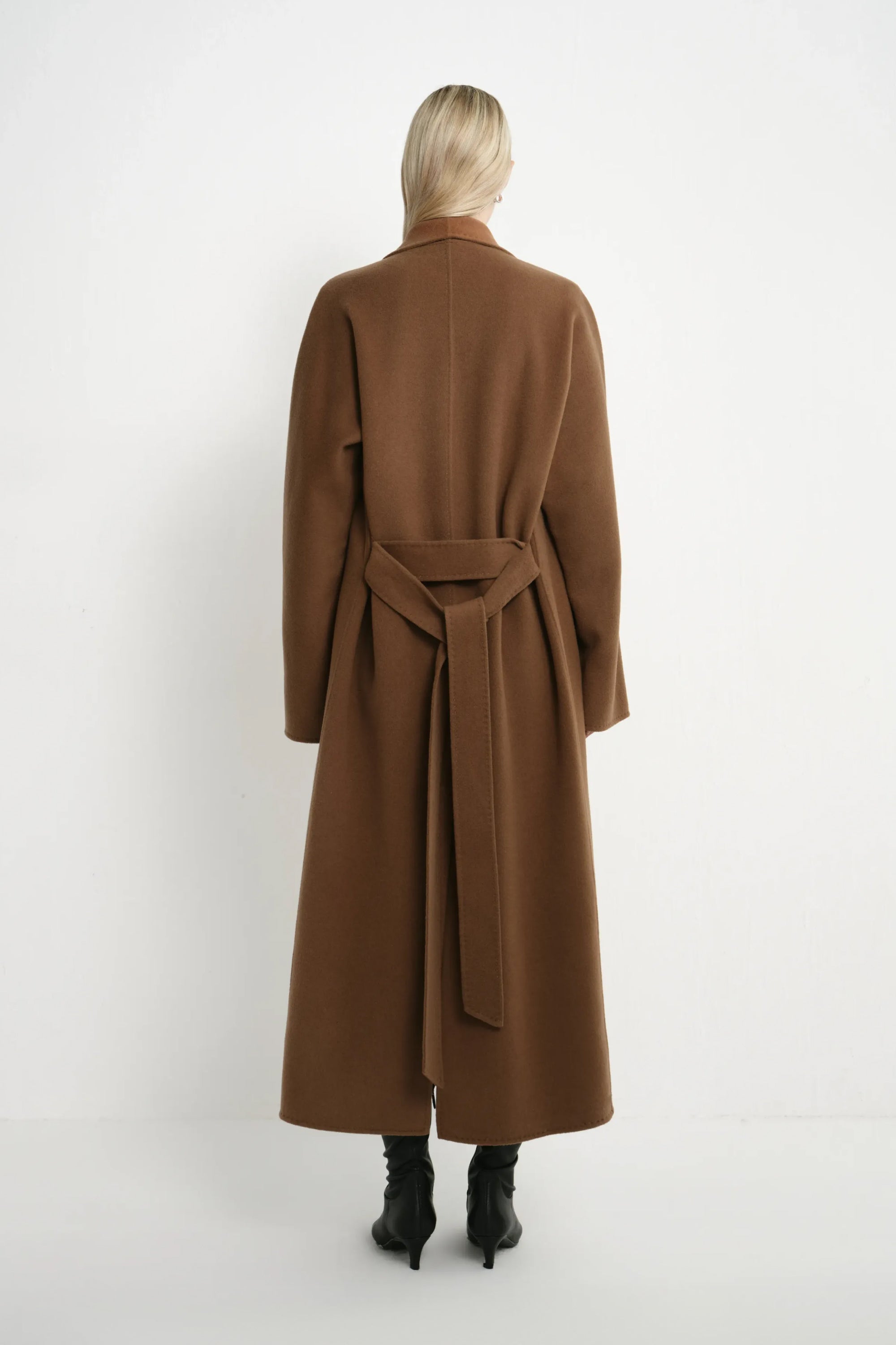 A model viewed from the back wearing a long brown belted coat and black ankle boots against a plain white studio background.