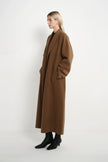 A woman stands in profile wearing a long, oversized brown coat over a black outfit with black ankle boots, photographed against a plain light gray studio background.