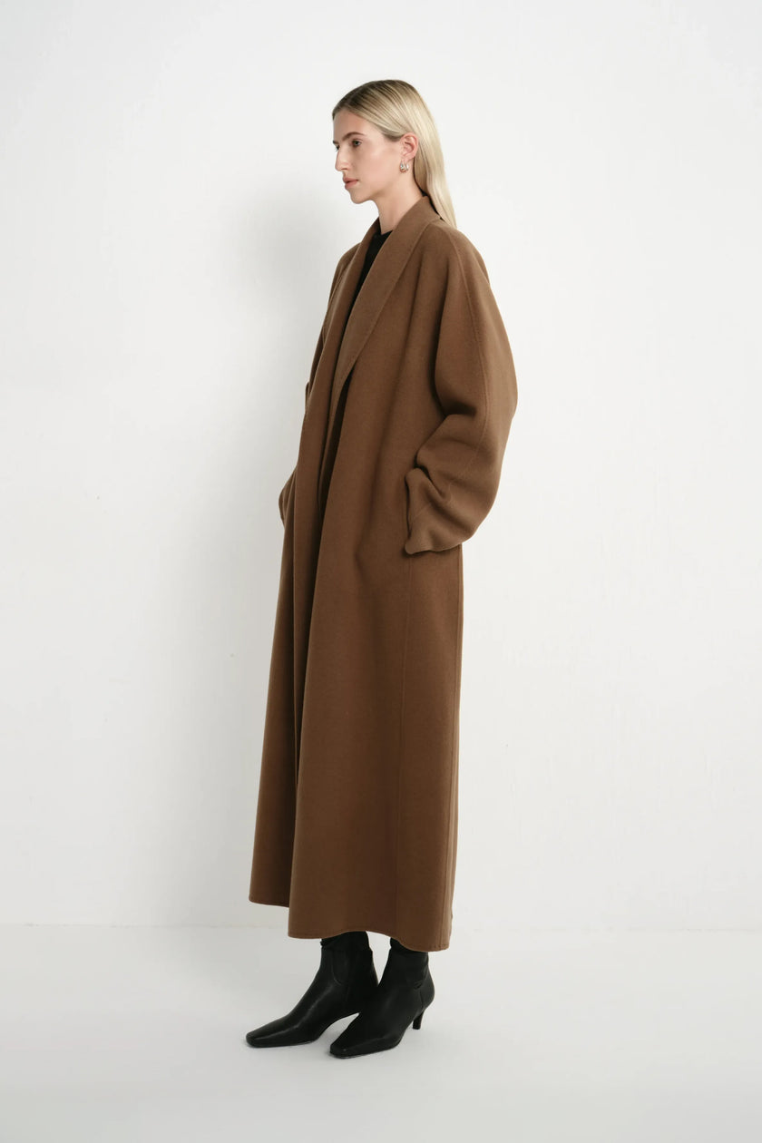 A woman stands in profile wearing a long, oversized brown coat over a black outfit with black ankle boots, photographed against a plain light gray studio background.