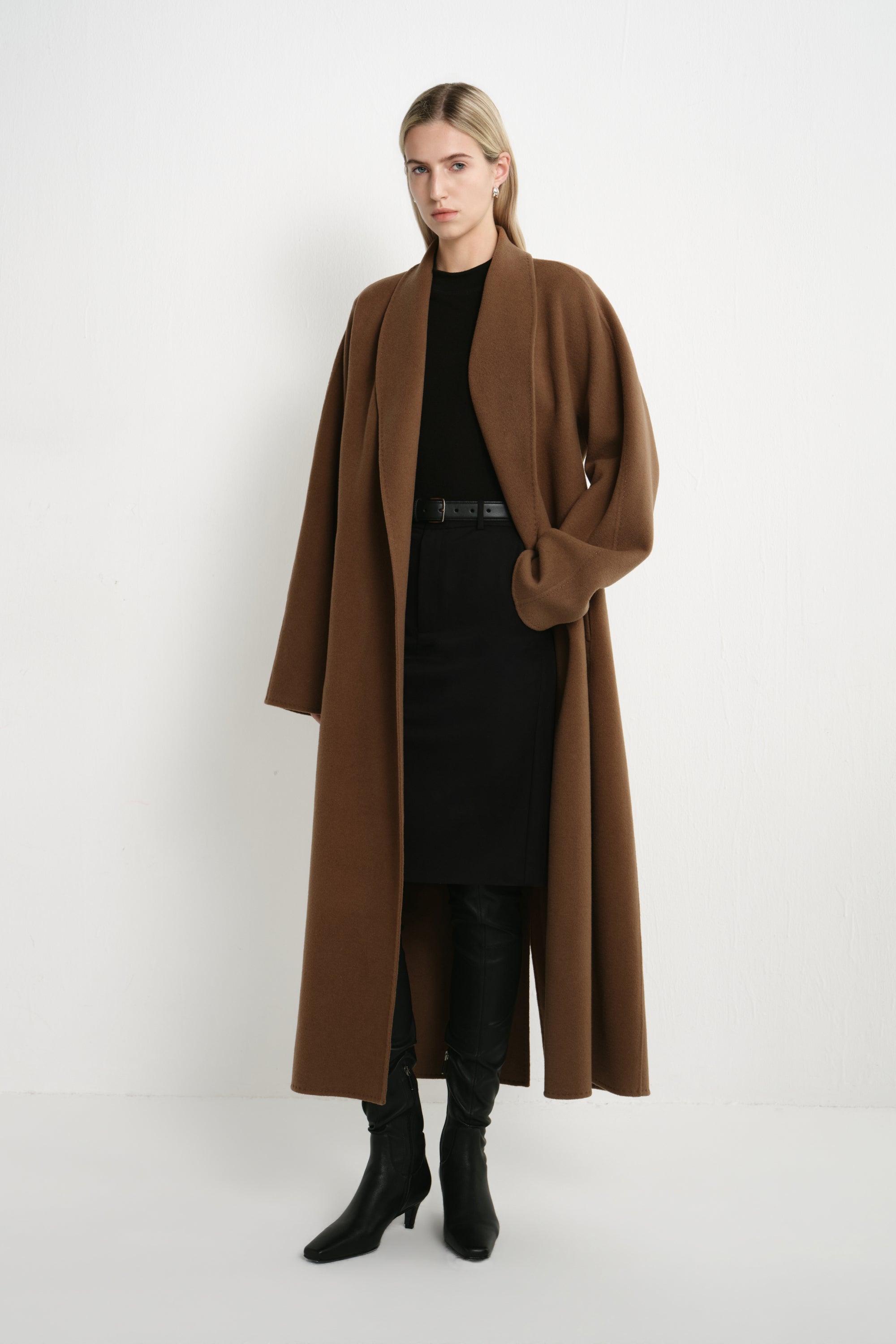 A woman modeling a long brown open-front coat over a black dress and black boots, standing against a plain white studio background.