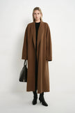 A model stands against a plain white studio background wearing a long, open-front camel brown coat over a black top and black ankle boots, while holding a black handbag.