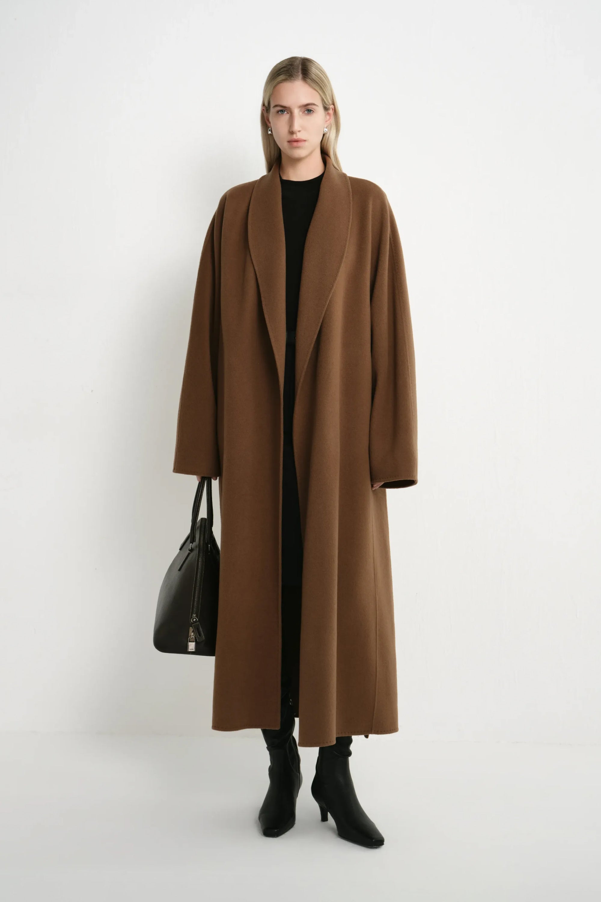 A model stands against a plain white studio background wearing a long, open-front camel brown coat over a black top and black ankle boots, while holding a black handbag.
