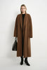 A model stands against a plain white studio background wearing a long, open-front camel brown coat over a black top and black ankle boots, while holding a black handbag.