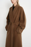 A woman wearing a long camel-brown belted coat over a black top, photographed against a plain light background.