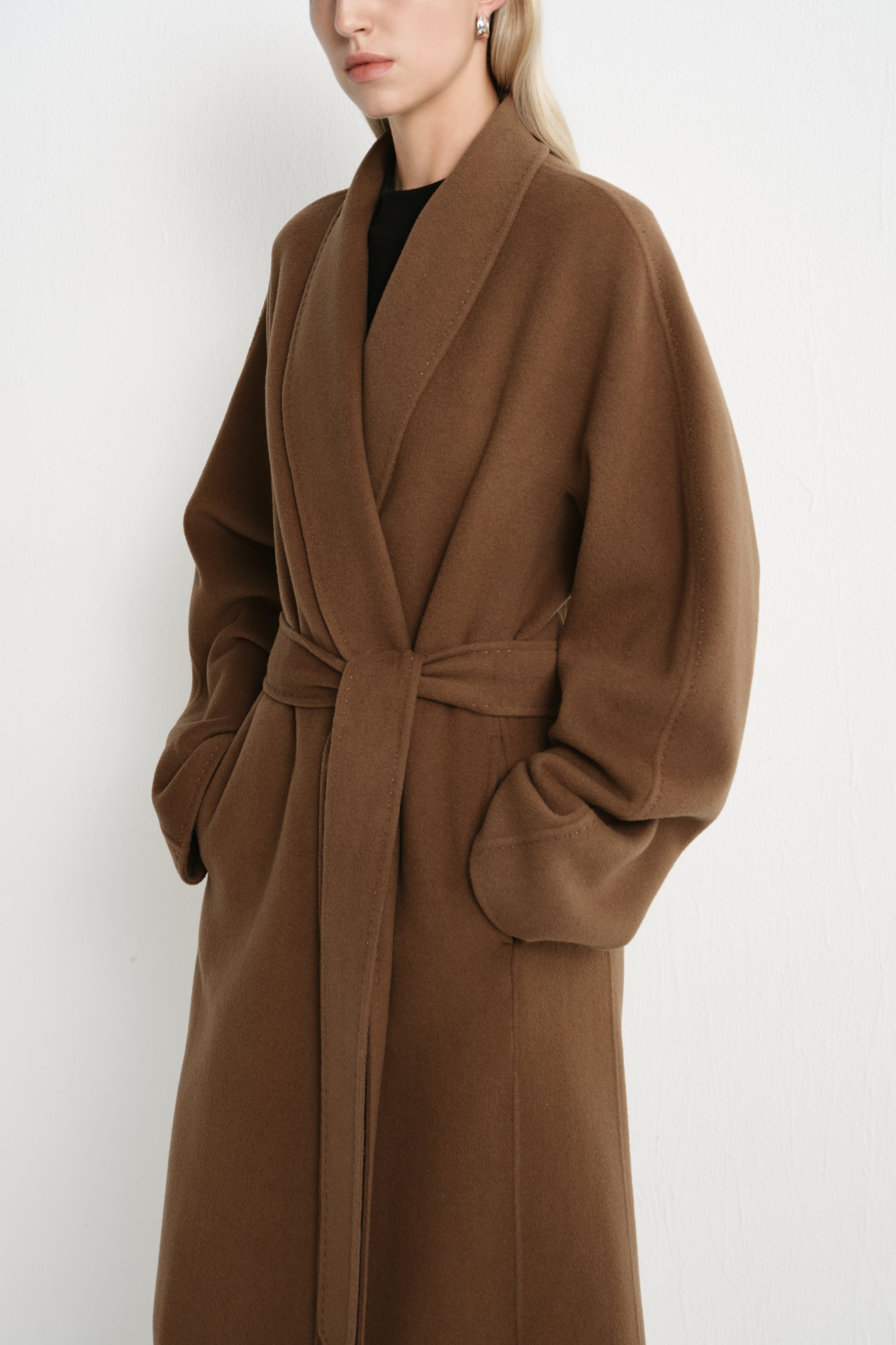 A woman wearing a long camel-brown belted coat over a black top, photographed against a plain light background.