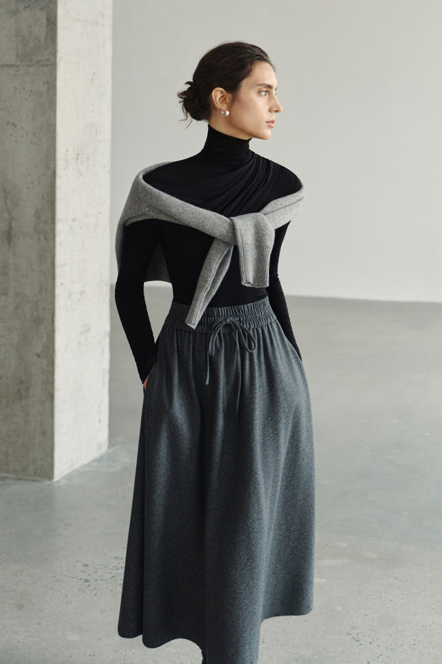 Three-quarter view of a model in a black turtleneck top with a grey sweater wrapped across the shoulders, paired with a charcoal drawstring maxi skirt.