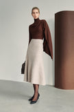 Editorial view of a model wearing a dark brown asymmetric draped turtleneck top with a light beige A-line midi skirt, sheer tights and black pointed pumps.