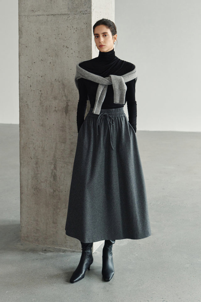 A model wearing a black turtleneck top with a grey sweater draped over the shoulders, styled with a charcoal drawstring maxi skirt and black ankle boots.