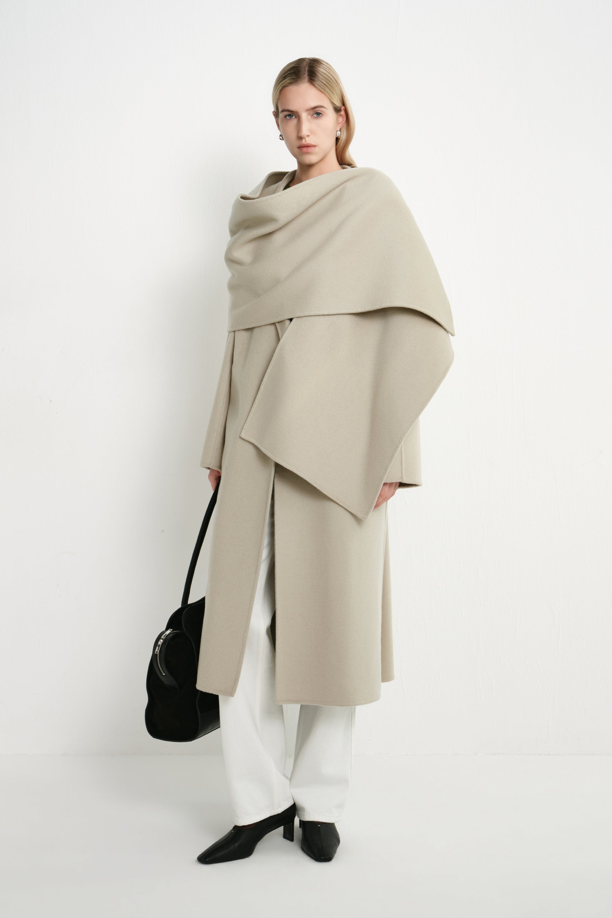 A model stands against a plain white studio background wearing a draped, oversized light beige wrap coat over white wide-leg pants and black pointed shoes, while carrying a black handbag.