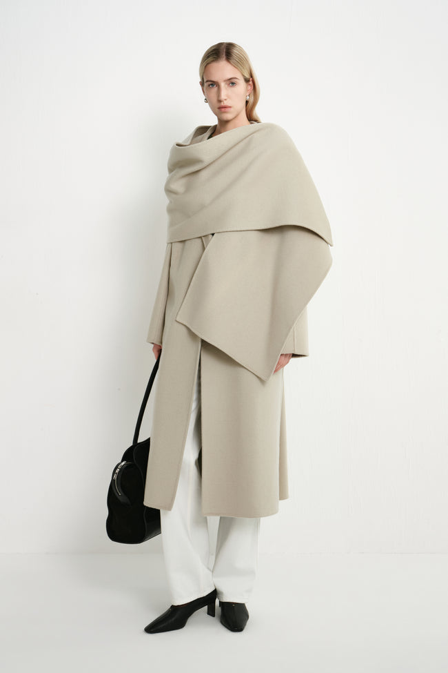 A model stands against a plain white studio background wearing a draped, oversized light beige wrap coat over white wide-leg pants and black pointed shoes, while carrying a black handbag.