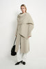 A model stands against a plain white studio background wearing a draped, oversized light beige wrap coat over white wide-leg pants and black pointed shoes, while carrying a black handbag.