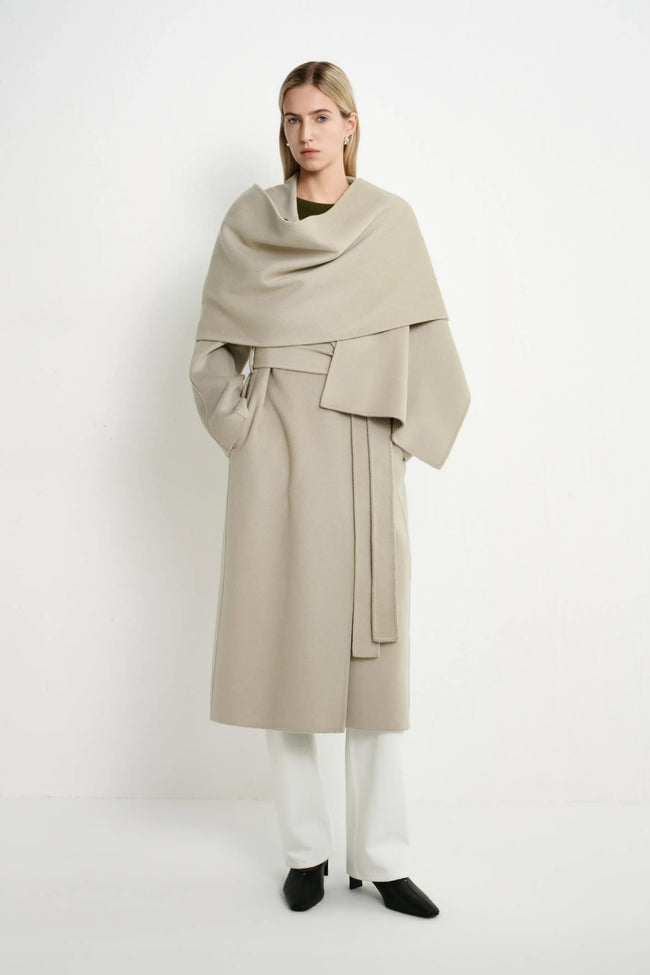 A model stands against a plain white studio background wearing a long taupe-beige wrap coat with a draped shawl, white wide-leg trousers, and black pointed shoes.