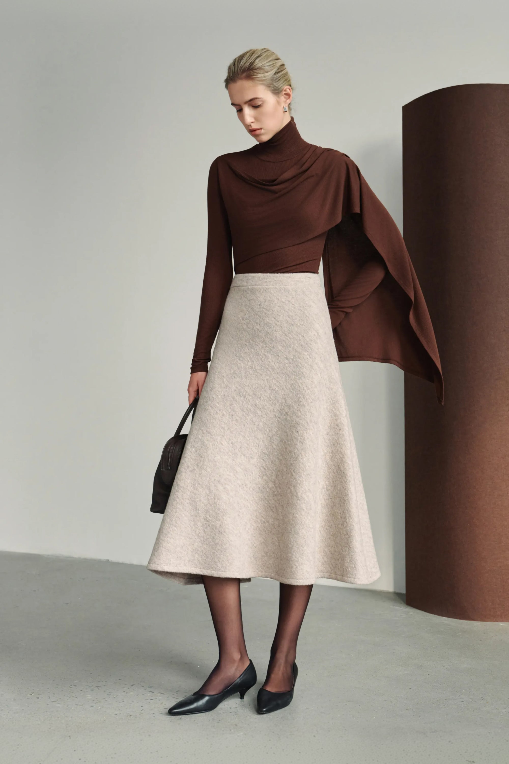 A model wearing a dark brown draped turtleneck top with an extended asymmetric shoulder panel, styled with a light beige A-line midi skirt and black pointed pumps.