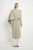 A model viewed from the back wearing a light beige draped wrap coat over white trousers, set against a plain white studio background.