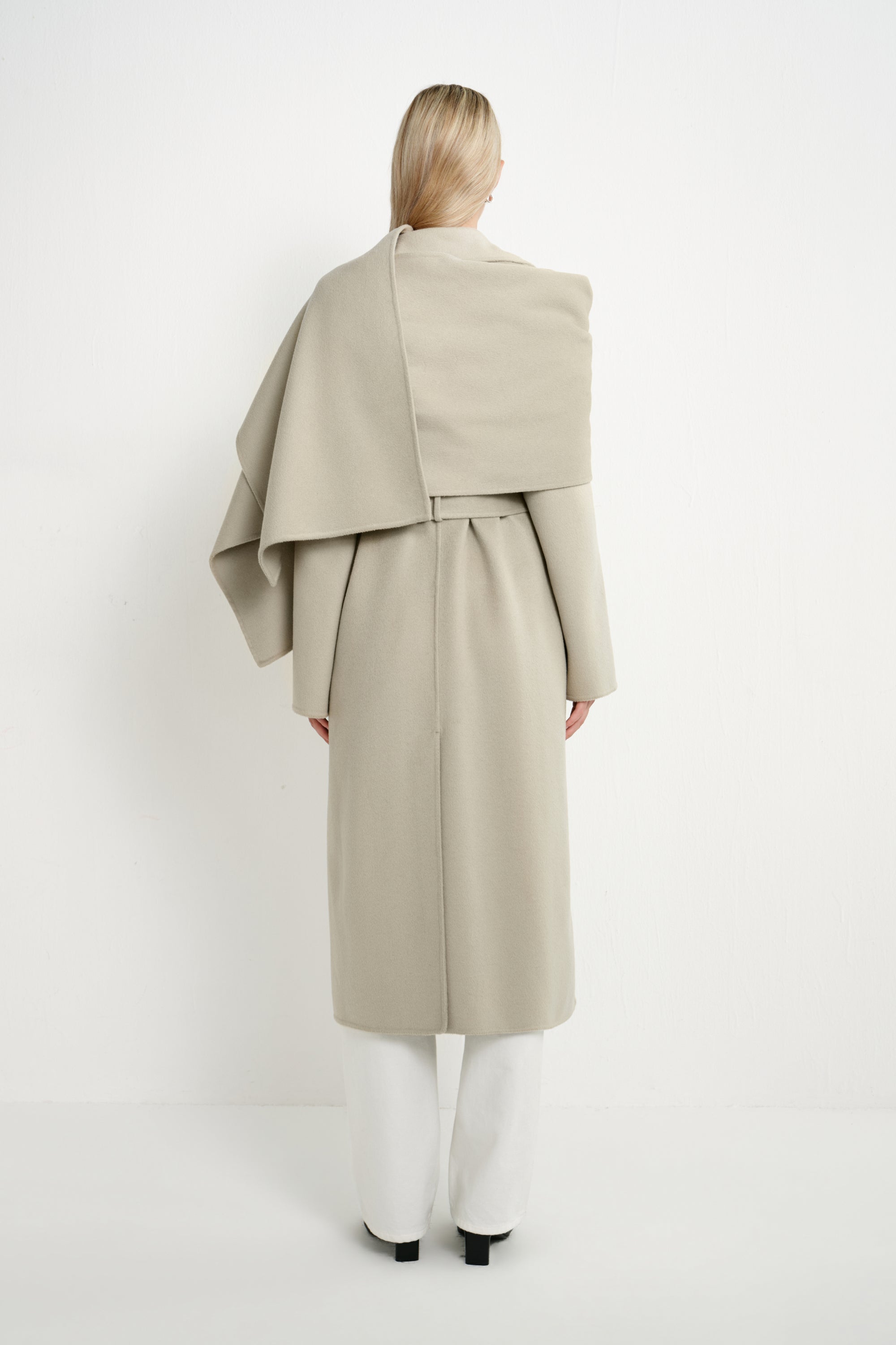 A model viewed from the back wearing a light beige draped wrap coat over white trousers, set against a plain white studio background.