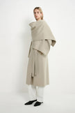 A model wearing a long, draped beige coat with a wide shawl collar, white wide-leg trousers, and black pointed shoes, photographed against a clean white studio background.