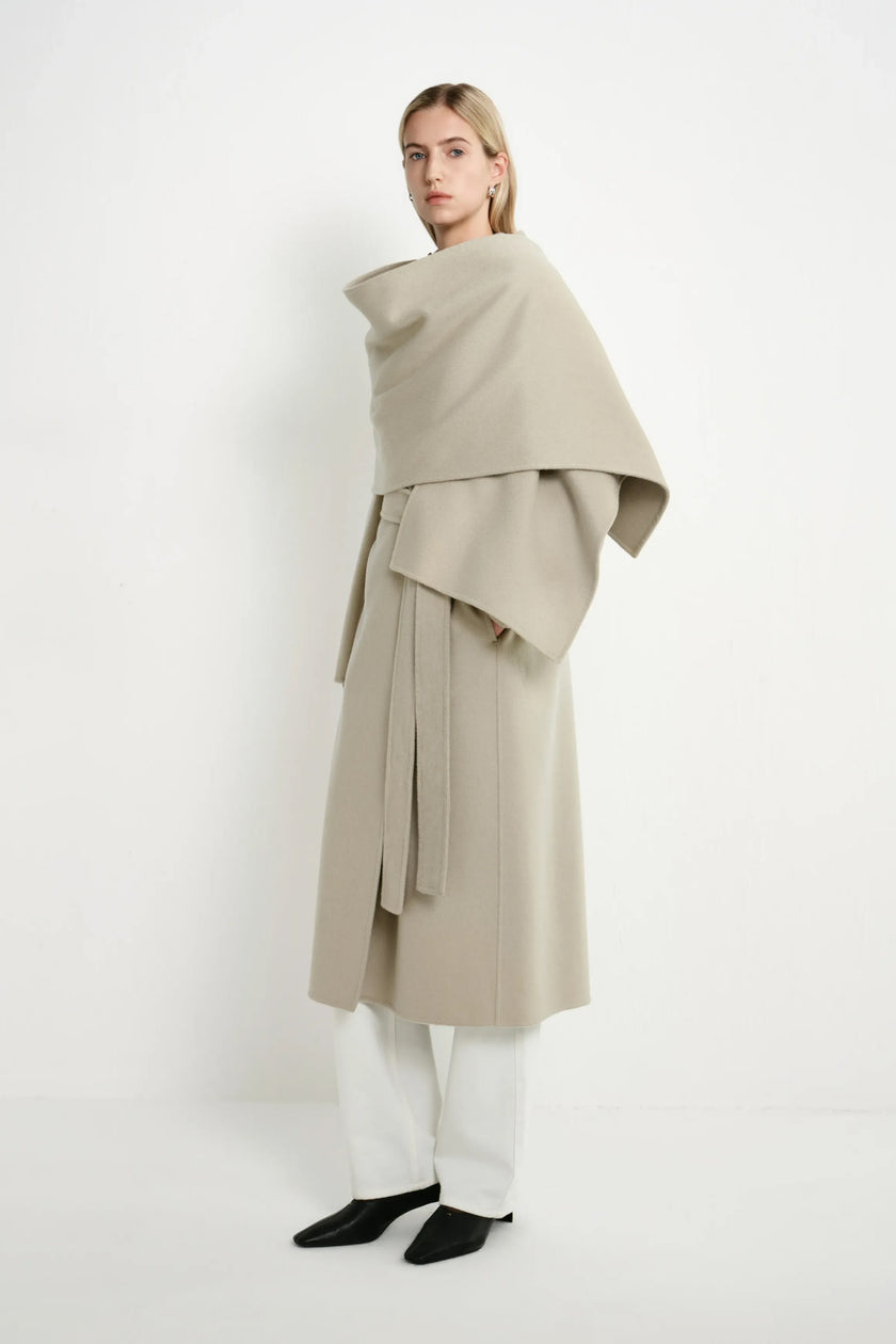 A model wearing a long, draped beige coat with a wide shawl collar, white wide-leg trousers, and black pointed shoes, photographed against a clean white studio background.