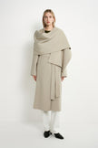 A model stands against a plain white studio background wearing a long, draped beige coat with a wide shawl-like collar, white pants, and black shoes.
