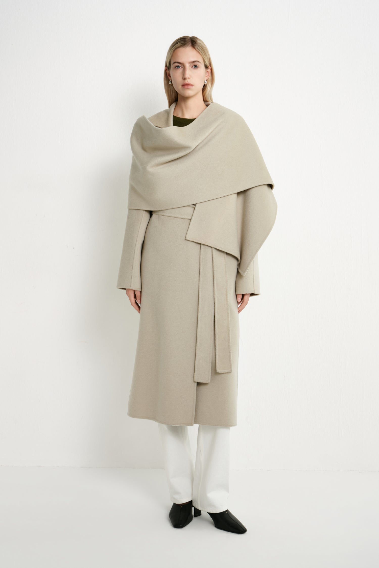 A model stands against a plain white studio background wearing a long, draped beige coat with a wide shawl-like collar, white pants, and black shoes.